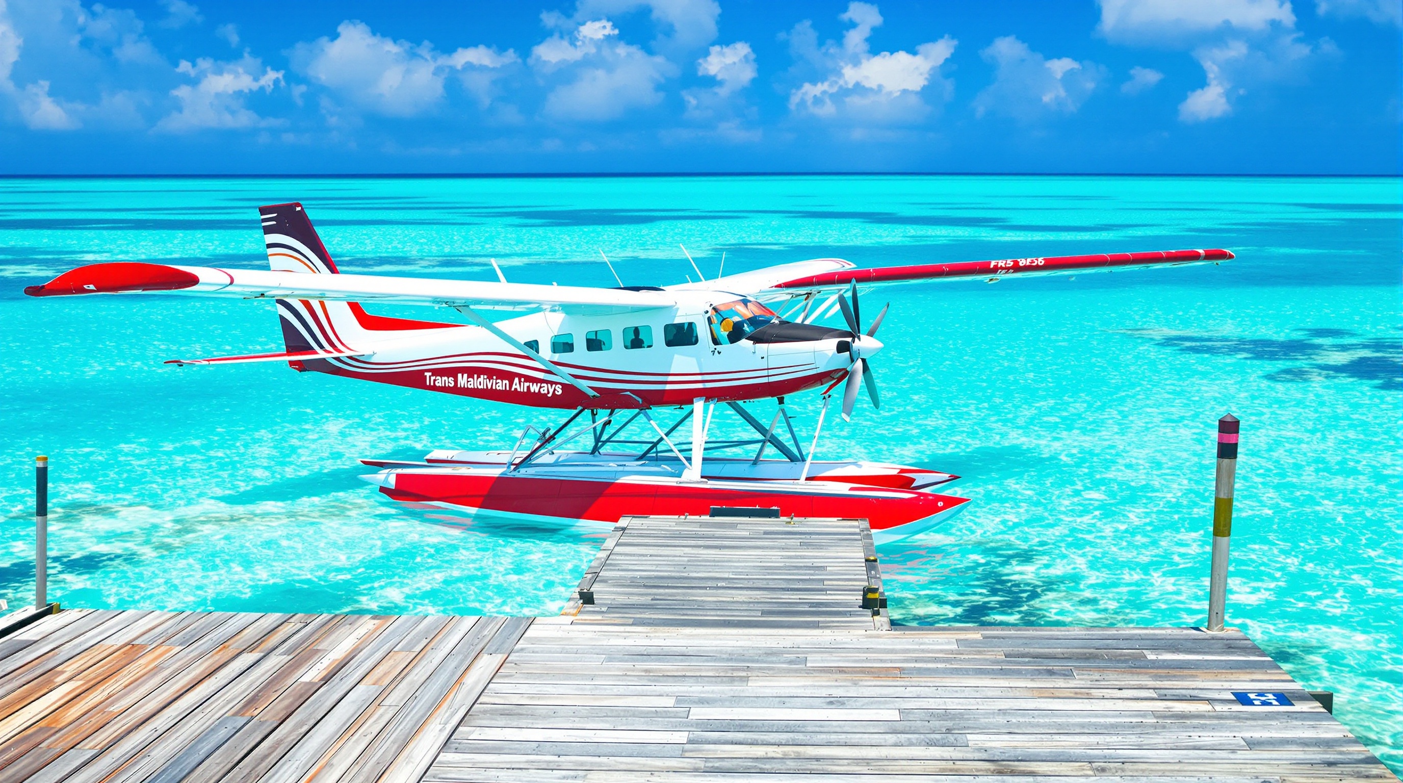 Hydravion Trans Maldivian Airways rouge et blanc amarré à un ponton en bois devant un lagon turquoise.